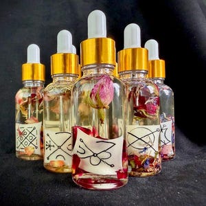 Könnte beinhalten: Eine Sammlung klarer Glasflaschen mit goldfarbenen Kappen und weißen Tropfern. Jede Flasche enthält ein Blumenarrangement aus getrockneten Blütenblättern und ein Etikett mit schwarzer Linienkunst. Die Flaschen sind auf einer schwarzen Oberfläche angeordnet.