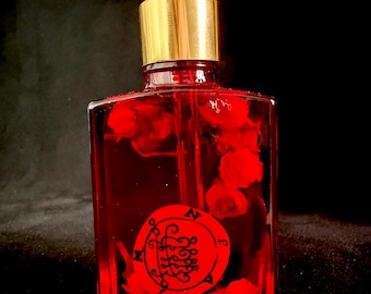 Op de afbeelding: Een vierkante glazen fles gevuld met een dieprode vloeistof en kleine rode rozenknopjes. De fles heeft een goudkleurige hals, een zwarte druppelaar en een rond rood etiket aan de voorzijde met een zwart, ingewikkeld, wervelend zegelontwerp.