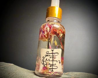 Op de afbeelding: Een heldere glazen fles met een gouden dop en druppelaar, gevuld met een heldere vloeistof en gedroogde bloemen. Een wit etiket met zwart schrift is op de fles bevestigd. De fles staat op een grijs stenen oppervlak.