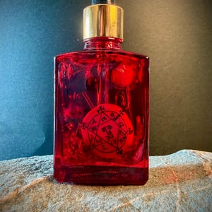 Pode incluir: Uma garrafa de vidro vermelho com tampa dourada e conta-gotas preto. A garrafa contém um líquido vermelho e um emblema circular vermelho com símbolos pretos. A garrafa está sobre uma superfície de pedra.