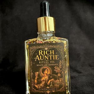 May include: A clear glass dropper bottle filled with yellow oil and dried botanicals. The black label features gold text reading Rich Auntie Ritual Oil Wealth and Abundance Witch Elixir and an illustration of a person holding a goblet.