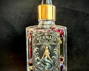 Op de afbeelding: Een rechthoekige, doorzichtige glazen fles met een gouden druppeldop, gevuld met olie en gedroogde bloemblaadjes. Het etiket toont een illustratie van een zeemeermin met de tekst MERMAID RITUAL OIL, BEAUTY, GLAMOUR, ATTRACTION en WITCH ELIXIR.