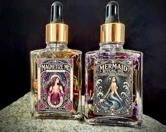 Op de afbeelding: Twee doorzichtige glazen druppelflesjes met gouden doppen gevuld met botanische olie. Het linker etiket bevat de tekst MAGNETIZE ME ATTRACTION ELIXIR met een illustratie van een vrouw. Het rechter etiket toont MERMAID RITUAL OIL WITCH ELIXIR met een zeemeermin.