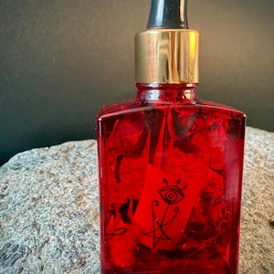 Puede incluir: Una botella de vidrio rojo con un cuentagotas negro y un collar dorado. Dentro de la botella hay una tarjeta roja con un símbolo de ojo negro y otras marcas. La botella está sobre una superficie de piedra gris.