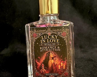 May include: A clear glass dropper bottle with a gold cap containing a liquid with visible botanical pieces. The label features the text LUCKY IN LOVE RITUAL OIL ROMANCE & ATTRACTION WITCH ELIXIR and an illustration of a person with long dark hair.