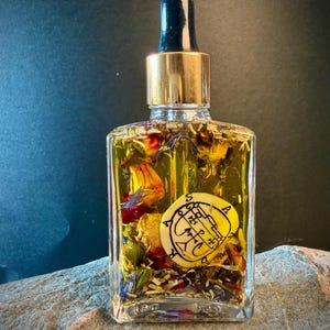 Könnte beinhalten: Eine klare Glasflasche gefüllt mit gelber Flüssigkeit und getrockneten Blumen, gekrönt von einer goldenen und schwarzen Pipette. Ein rundes, gelbes Emblem mit schwarzen Symbolen ist in der Flasche sichtbar. Die Flasche steht auf einer grauen Steinoberfläche.