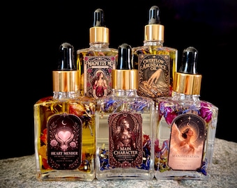 Op de afbeelding: Vijf rechthoekige glazen druppelflesjes met gouden doppen, gevuld met botanische oliën en gedroogde bloemen. De etiketten bevatten illustraties en de tekst: Heart Mender, Magnetize Me, Overflow Abundance Oil, Main Character Confidence Oil en Manifestation Oil.