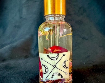 Op de afbeelding: Een doorzichtige glazen druppelfles met een gouden kraag en een witte druppeltop. De fles bevat een transparante vloeistof met diverse kleurrijke bloemblaadjes erin. Een wit rechthoekig etiket toont een zwarte lijntekening van vier maansikkels in een kruispatroon rond een centraal punt.