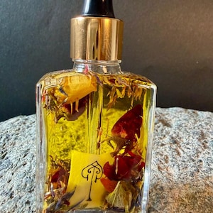 Può includere: Una bottiglia di vetro trasparente riempita di liquido giallo e fiori secchi, erbe e petali rossi. La bottiglia ha un tappo dorato e un contagocce nero. Un'etichetta gialla con un'illustrazione nera è visibile all'interno.