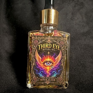 Può includere: Un flacone in vetro trasparente con tappo contagocce dorato contenente olio e petali di fiori secchi. L'etichetta riporta il testo THIRD EYE RITUAL OIL e WITCH ELIXIR con un grafico di un occhio alato circondato da filigrana dorata.