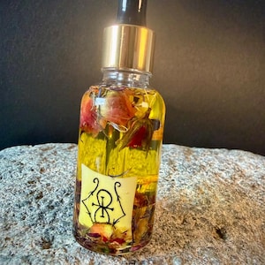 May include: A clear glass bottle containing yellow liquid and dried flowers, with a gold and black dropper. A yellow label with a black symbol is attached. The bottle is placed on a grey stone surface.