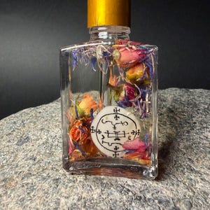 Op de afbeelding: Een heldere, rechthoekige glazen fles met een gouden dop en witte druppelaar. De fles bevat gedroogde bloemen in roze, oranje en blauwe tinten, en een wit cirkelvormig embleem met zwarte symbolen. De fles staat op een grijze stenen ondergrond.