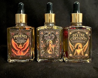 May include: Three rectangular glass dropper bottles with gold caps, each containing a clear liquid and botanical elements. The labels feature ornate borders and text: Phoenix The Comeback Rebirth, That Girl Glow Up, and Divine Certainty Manifestation Oil.