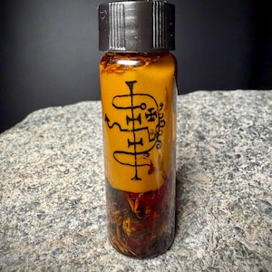 Może przedstawiać: Mała szklana butelka z czarną zakrętką, wypełniona żółtym płynem i suszonymi ziołami. Butelka ma czarny symbol z przodu. Butelka stoi na szarej kamiennej powierzchni.