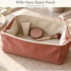 May include: A wide-open zipper pouch in a dusty rose color with a cream-colored interior featuring thin vertical stripes. The pouch contains a small squeeze tube, a round compact, and a makeup brush. The text reads Wide-Open Zipper Pouch.