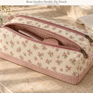 Può includere: Un astuccio rettangolare con doppia cerniera in tessuto color crema con stampa di rose rosa e bordo a quadretti vichy rosa e bianchi. L'astuccio ha due scomparti con cerniera, uno dei quali è aperto. Il testo recita Rose Garden Double Zip Pouch.