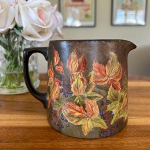 May include: A brown ceramic jug with a handle, featuring a painted design of autumn leaves and berries.