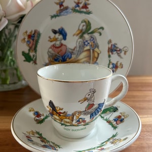 May include: A vintage teacup and saucer set featuring a charming design of ducks. The cup and saucer are white with a gold trim and depict a colourful illustration of ducks in a boat with the text "Dizzy Ducklings".