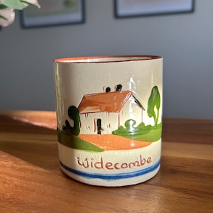 May include: A cylindrical ceramic mug with a cream-colored background. The mug features a painted scene of a house with an orange roof, green trees, and the word "Widecombe" in red. A blue stripe encircles the base.