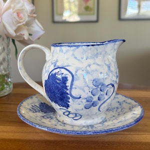 May include: A white ceramic milk jug with a blue and white floral design. The jug has a handle and a spout. It is sitting on a matching saucer.