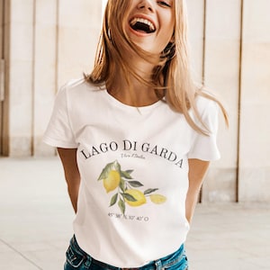 Puede incluir: Camiseta blanca con la inscripción "LAGO DI GARDA" y una ilustración de limón. La camiseta también incluye las coordenadas 45° 38′ N, 10° 40′ O. La modelo lleva vaqueros azules.