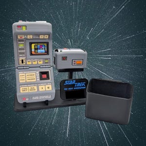 May include: A grey Star Trek The Next Generation TR-590 Tricorder VII replica featuring a hinged design, a digital screen, and various control buttons. It is displayed on a black stand with a matching black case and a Star Trek logo plaque.