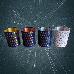 May include: Four cylindrical candle holders with geometric cutouts. The holders are navy blue, with gold, copper, silver, and white interiors. The cutouts create a repeating pattern of small triangles. The background is dark with white streaks.