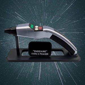May include: A silver and black Type II Phaser prop from "Endgame" on a black stand. The prop has a curved handle with a ribbed grip, a black nozzle, and a control panel with green and red buttons. The text "ENDGAME" TYPE II PHASER is on a black plaque.