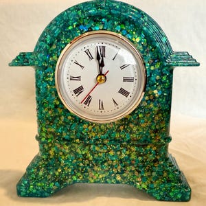 May include: A decorative clock with a teal and gold glitter finish. The clock face is white with black Roman numerals and gold trim. The hands are black and red. The clock has an arched top and a rectangular base.