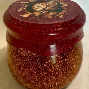 May include: A decorative jar with a red glitter lid featuring a cat illustration. The jar's body is filled with gold glitter, creating a sparkling effect. The lid has a glossy finish.