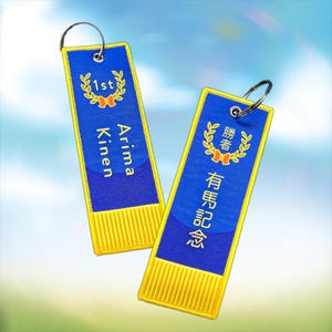 May include: Two blue and gold keyrings with silver hoops. One has the text "1st" and "Arima Kinen" in white. The other has Japanese text and a laurel wreath design. Both have gold fringe.