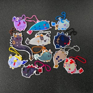 Pode incluir: Uma coleção de doze porta-chaves de acrílico com vários designs de criaturas estilizadas em diversas cores, incluindo roxo, azul, branco, preto e azul-petróleo. Cada pendente está preso a uma pequena corrente de bolas colorida.