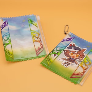 May include: Two clear, rectangular pouches with a colorful design. One pouch features a cartoon character sticker. The pouches have a green, scalloped edge and a zipper closure. The background of the pouches is a sky and grass scene.