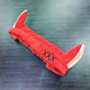 Puede incluir: Una diadema de felpa roja con detalles en forma de oreja en posición vertical con forro blanco. La diadema tiene las letras bordadas "XXX" en negro en un lado. El artículo está diseñado para ser usado en la cabeza.