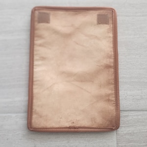 Puede incluir: Una almohadilla rectangular de color beige con un borde marrón y dos pequeños parches cuadrados de velcro. La almohadilla tiene un aspecto ligeramente arrugado y está sobre un fondo gris claro.