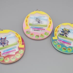 May include: Three colourful, round, handheld digital pet toys featuring cartoon character graphics. Each toy has a screen displaying a character, surrounded by a yellow and pink or blue frame. The toys have buttons and a keychain attachment.