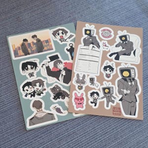 Puede incluir: Dos hojas de pegatinas rectangulares con varias ilustraciones estilo anime. Una hoja es de color verde salvia con personajes, un tigre y una escena urbana. La otra es de color tostado con personajes con cabezas de televisión y el texto SYR WAS HERE.