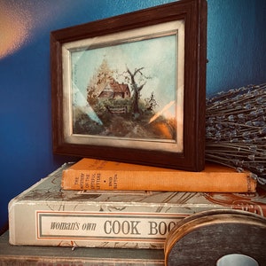 May include: A stack of vintage books with a framed watercolor painting of a cottage and a tree on top. The top book is orange and says "The Mystery of the Spiteful Letters" by Enid Blyton. The middle book is white and says "Woman's Own Cook Book". The bottom book is blue and has text in a foreign language. A wooden spool with a white center is in the foreground.