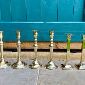 Peut inclure: Six bougeoirs en laiton de différentes formes sont alignés. Les bougeoirs présentent des bases et des tiges variées, avec une finition polie. En arrière-plan, une porte en bois turquoise et une surface en pierre.