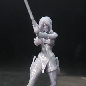 May include: A gray 3D printed miniature figurine of a female warrior holding a sword. The figurine is standing on a round base.