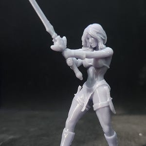 May include: A gray 3D printed miniature figurine of a female warrior holding a sword in a fighting pose. The figurine is standing on a round base.