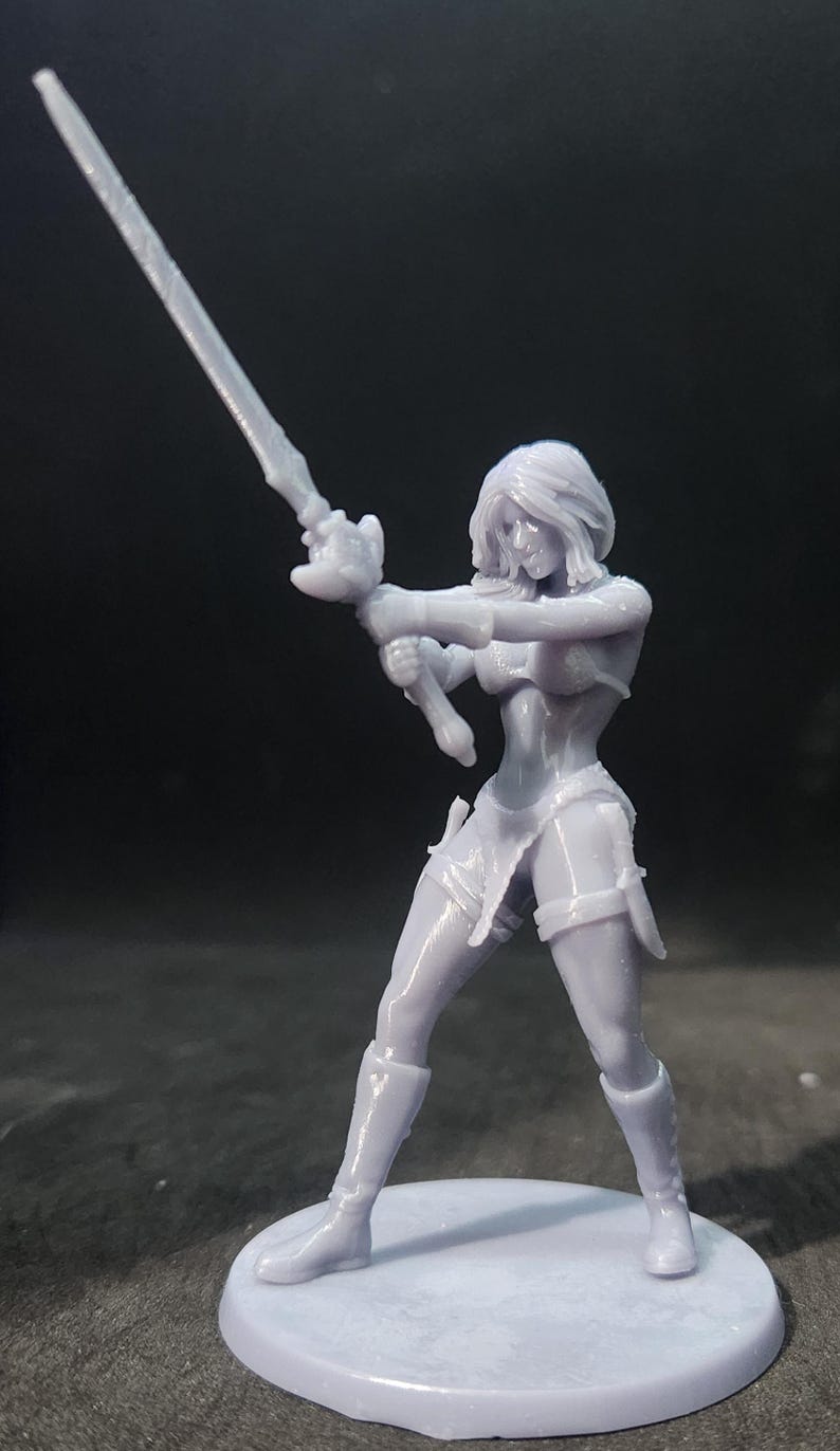 May include: A gray 3D printed miniature figurine of a female warrior holding a sword in a fighting pose. The figurine is standing on a round base.