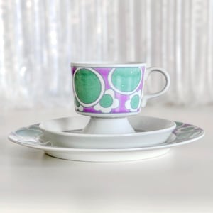 Puede incluir: Un juego de taza y platillo de té vintage. La taza presenta un diseño floral púrpura y verde. El platillo y el plato son blancos con un patrón floral a juego. El juego se exhibe sobre una superficie blanca con un fondo suave y borroso.