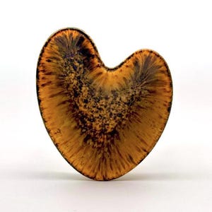 Puede incluir: Un objeto en forma de corazón con un degradado de tonos ámbar cálido y amarillo dorado que transicionan hacia un centro marrón oscuro y moteado. La superficie muestra una textura radial y fibrosa que se extiende desde el centro al borde.
