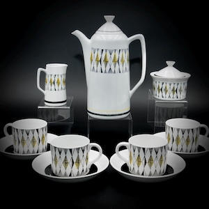 May include: A white ceramic tea set with a geometric pattern in black and gold. The set includes a teapot, creamer, sugar bowl, and four cups and saucers.