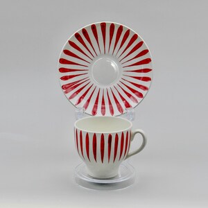 May include: A white teacup and saucer set with a red stripe design. The cup and saucer are made of ceramic and have a glossy finish. The saucer has a radial pattern of red stripes, while the cup has vertical red stripes.