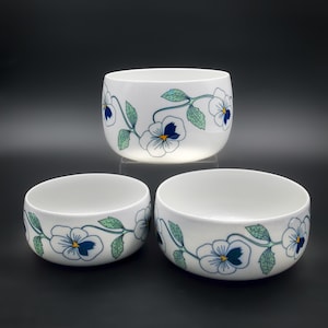 May include: Three white ceramic bowls with a floral design. The bowls have a rounded shape and are decorated with a pattern of blue and white pansies with green leaves. The bowls are of different sizes and are arranged on a dark surface.