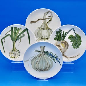 May include: Four white ceramic plates with hand-painted vegetable designs. The plates feature illustrations of garlic, onions, and other root vegetables in shades of green, brown, and white. The plates are displayed on clear stands against a blue background.