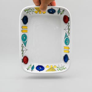 Puede incluir: Plato rectangular de cerámica blanca con un borde colorido. El borde presenta ilustraciones dibujadas a mano en azul, amarillo, rojo y verde. El plato está sostenido por una mano.