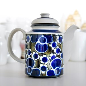 Könnte beinhalten: Keramik-Teekanne mit blau-weißem Blumenmuster. Die Teekanne hat einen weißen Ausguss und Henkel sowie einen grauen Deckel. Das Design zeigt stilisierte Blumen auf dunklem Hintergrund.